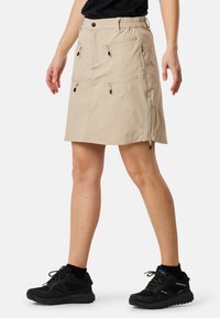 Beige outdoor skirt with elastic waistband, multiple zippered pockets, knee-length, and synthetic fabric for durability. Paired with black sneakers.