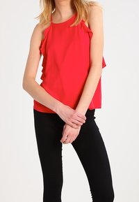 Top sin mangas de color rojo con detalles con volantes, tejido ligero y un corte holgado. Se lleva con pantalones ajustados negros. Diseño de escote sencillo.