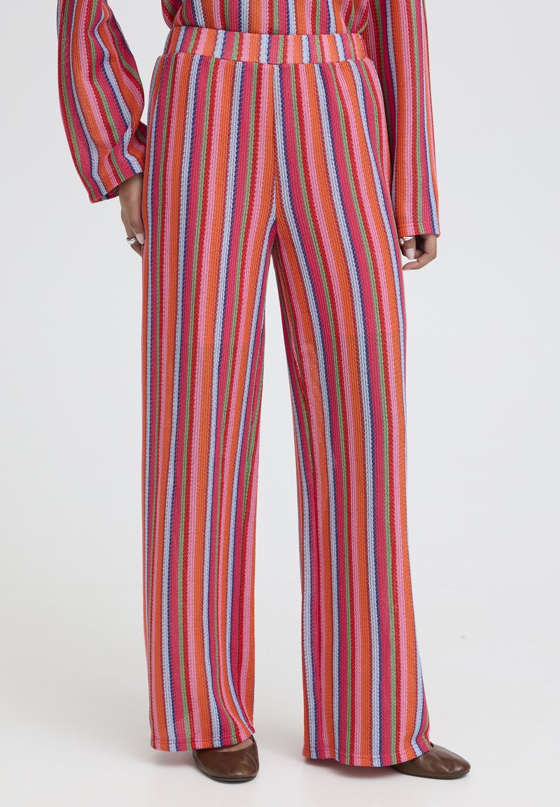 Personne portant un pantalon taille haute à jambes larges avec des rayures verticales multicolores, assorti à des chaussures marron à bouts pointus et un haut rayé assorti.