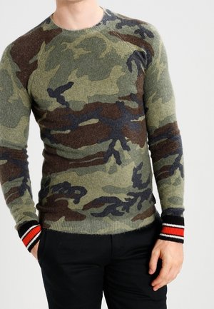 Homme portant un pull à motif camouflage vert et marron avec des poignets rayés rouges, blancs et noirs, associé à un pantalon noir.
