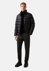 Černá péřová bunda s vysokým límcem, zapínáním na zip a prošívaným designem, spárovaná s tmavými kalhotami a černými šněrovacími teniskami.