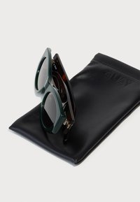 Ochelari de soare cu rame verzi și cu model țestoasă, având lentile închise la culoare, așezați pe un toc din piele neagră timbrat cu numele brandului "QUAY."