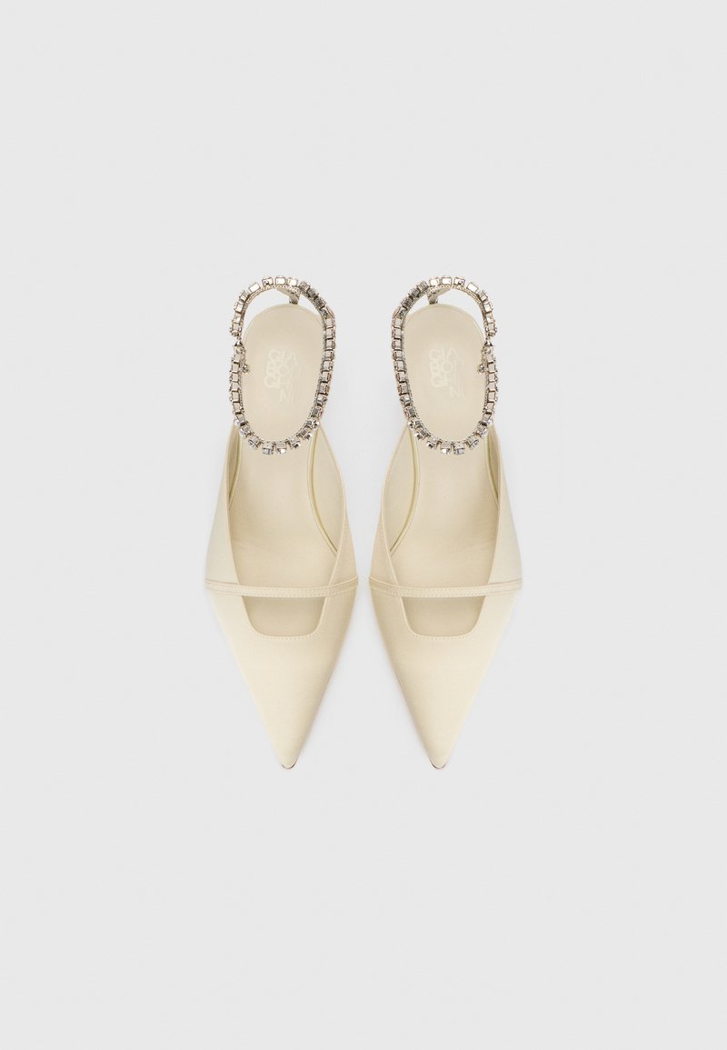 Creme spidse slingback flade sko med en tynd rem over foden og rhinestone-dekorerede ankelremme på en hvid baggrund.
