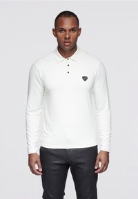 Weißes langärmeliges Polohemd mit klassischem Kragen und drei schwarzen Knöpfen, versehen mit einem schwarzen Logopatch auf der Brust.