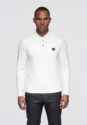 Vit långärmad pikétröja med klassisk krage och tre svarta knappar, med en svart logotypmärke på bröstet.