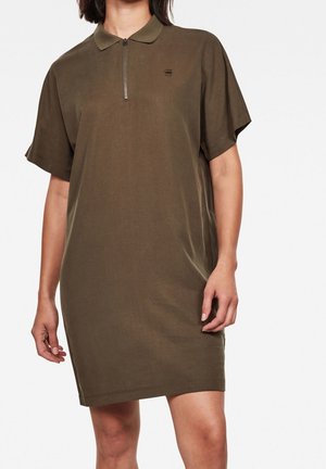 Frau trägt ein olivgrünes Poloshirt-Kleid mit kurzen Ärmeln, Frontreißverschluss und Kragen, stehend vor einem einfarbigen Hintergrund.