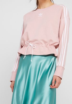 Kobieta w pastelowo różowej bluzie Adidas z białymi paskami i ściągaczem w talii, połączona z błyszczącą turkusową satynową spódnicą.