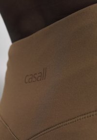 Gros plan sur un legging de sport marron taille haute avec le logo "casall" et des coutures visibles sur le tissu.