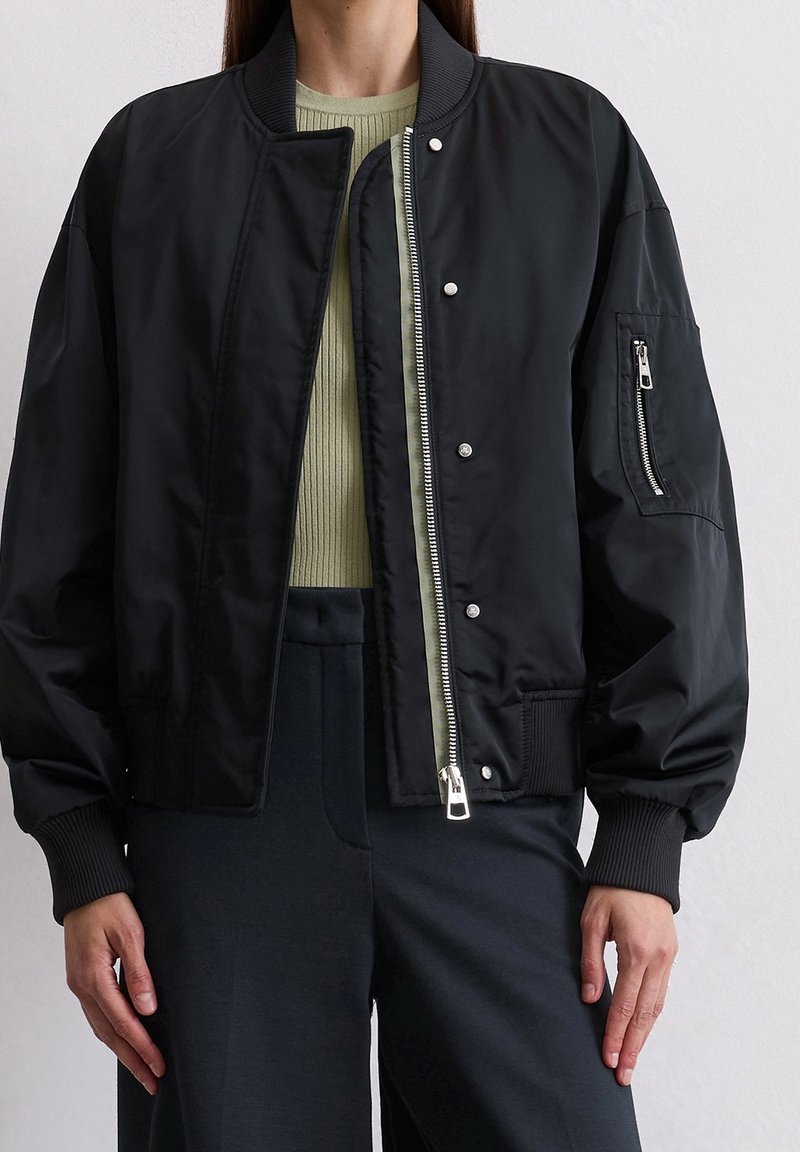 Femme portant une veste bomber noire avec poignets et col côtelés, partiellement zippée sur un top côtelé vert clair et un pantalon foncé.