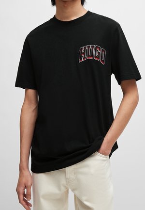 Person trägt ein schwarzes T-Shirt mit kurzen Ärmeln mit rotem und weißem "HUGO" Text auf der Brust und weiße Hose, Hand in der Tasche.