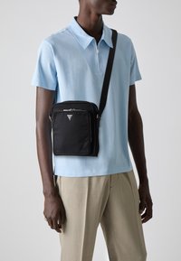 Borsa a tracolla in nylon nero con chiusura zip, dotata di un logo triangolare e di una tracolla rimovibile, indossata sopra una polo azzurra.