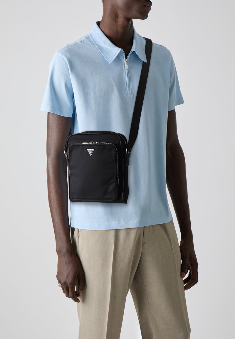 Borsa a tracolla in nylon nero con chiusura zip, dotata di un logo triangolare e di una tracolla rimovibile, indossata sopra una polo azzurra.