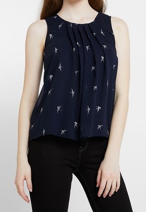 Blusa senza maniche blu navy con pieghe e piccoli motivi di uccellini bianchi, indossata con pantaloni neri.