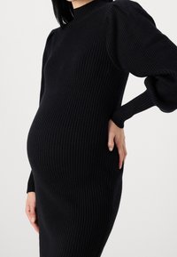 Robe noire moulante côtelée avec des manches longues et un col haut, conçue pour épouser l'abdomen, avec un tissu texturé et une silhouette ajustée.