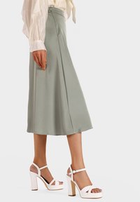 Gonna midi verde chiaro con una texture liscia e cerniera laterale, abbinata a sandali bianchi con tacco alto, dotati di cinturini incrociati e tacco spesso.