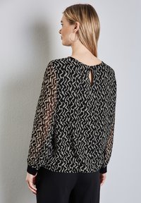 Schwarze und beige gemusterte Bluse mit durchsichtigen langen Ärmeln und einem Schlüsselaugen-Detail im Rücken; gerippte Bündchen am Handgelenk verleihen dem Design zusätzlichen Charme.
