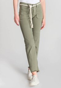 Pantalons en mélange de coton vert olive avec un ourlet effiloché et des poches cousues, maintenus par une ceinture en corde tressée. Portés avec des baskets blanches.