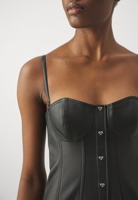 Femme portant un corset en cuir noir avec des bretelles fines et quatre boutons en forme de cœur argentés au centre devant.