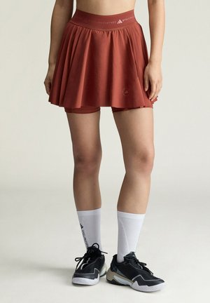 Personne portant une jupe de sport plissée couleur rouille avec des shorts intégrés, des chaussettes blanches mi-mollet et des chaussures de sport noires.