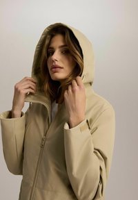 Beżowa kurtka z kapturem, z zamkiem błyskawicznym z przodu, luźny krój i regulowane mankiety. Gładka tekstura, z wysokim kołnierzem dla dodatkowej ochrony.