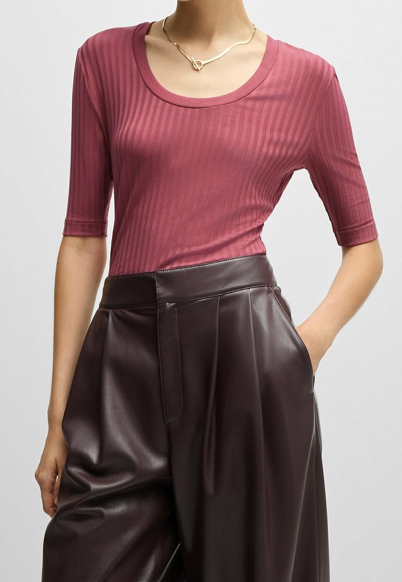 Femme portant un top à manches courtes côtelé, couleur rose poudré, et un pantalon en cuir marron foncé taille haute, main dans la poche.