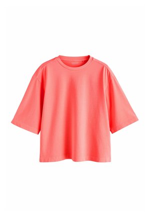 Vrouw met een los koraalkleurig shirt, denim bermudashorts en een roze geweven hoed staand op een stenen pier bij de oceaan.