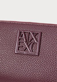 Textured burgundy leather wallet featuring a raised, glossy black Armani Exchange logo on the front, with a zip closure.
