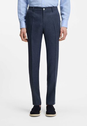 Mężczyzna ubrany w granatowe eleganckie spodnie, jasnoniebieską koszulę i czarne mokasyny z beżową podeszwą, stojący na białym tle.