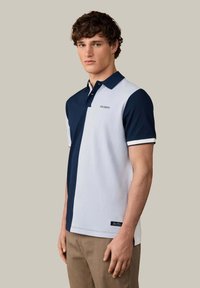 Joven con polo mitad azul marino, mitad blanco con el logo "Hackett", combinado con pantalones marrón claro, de pie frente a un fondo liso.