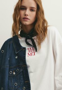 Jonge vrouw met golvend rood haar draagt een witte sweatshirt met rode tekst "THE MET", een zwarte halsdoek en een donker denimjack over één schouder gedrapeerd.