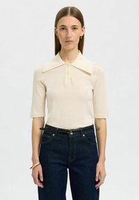 Camisa polo de manga corta color beige claro con un gran cuello, cierre de botones y textura acanalada, combinada con jeans de mezclilla oscura y un cinturón negro.