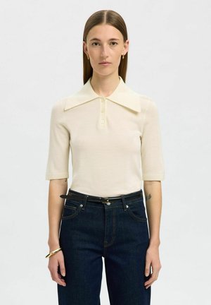 Polo shirt corto a maniche corte color beige chiaro con colletto ampio, apertura frontale con bottoni e texture a costine, abbinato a jeans in denim scuro e a una cintura nera.