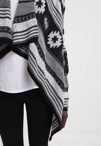 Persona con un chal tejido de patrón blanco y negro sobre una camisa blanca y pantalones negros, delante de un fondo claro y liso.