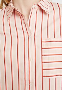 Camisa de botones rosa claro con rayas verticales en rojo y blanco, con un bolsillo en el pecho izquierdo y un diseño de cuello clásico.