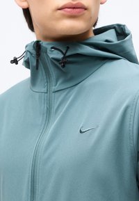 Κοντινό πλάνο ενός ατόμου που φοράει τυρκουάζ μπουφάν με κουκούλα, με μαύρο κορδόνι και λογότυπο Nike στο στήθος, το φερμουάρ είναι εντελώς κλειστό.