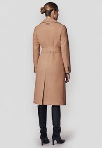 Długi, beżowy wełniany płaszcz z pasem w talii, szerokimi klapami i bocznymi rozcięciami. Noszony z czarnymi, sięgającymi kolan botkami o gładkiej fakturze.
