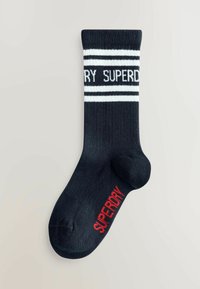Enkelt sort sok med hvide striber og delvist "SUPERDRY" tekst nær toppen, samt rød "SUPERDRY" tekst på svangen.
