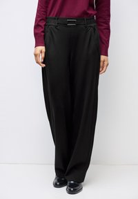 Femme portant un pantalon noir à jambes larges avec une ceinture noire et des chaussures noires, associée à un haut à manches longues de couleur rouge foncé, posant devant un fond blanc.