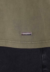 Olivenfarvet t-shirt med en glat tekstur, der har et sølvtag med teksten "Dropsize" nær kanten og er sat sammen med mørke jeans.