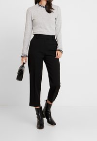 Grauer Langarmpullover mit einem gerüschten Kragen in Schwarz-Weiß kariert, kombiniert mit schwarzen schmalen Hosen und schwarzen Stiefeletten. Hält eine kleine Tasche.