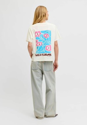 Kobieta z długimi blond włosami, ubrana w białą koszulkę z różowo-niebieskim kwiatowym wzorem i napisem „Wild Flower”, zestawioną z jasnoszarymi szerokimi dżinsami.