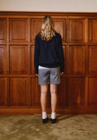 Ξανθός άνθρωπος με μαύρο πουλόβερ, καρό σορτς, λευκές κάλτσες και μαύρα παπούτσια, όρθιος μπροστά σε ξύλινο τοίχο, πάνω σε πράσινο χαλί.