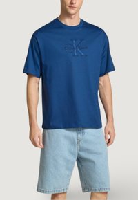 Mladý muž v modrém tričku s krátkými rukávy značky Calvin Klein a světlých modrých džínových šortkách, stojící na jednobarevném pozadí.