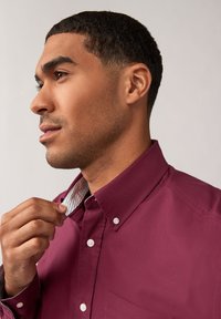 Camicia bordeaux con colletto a punta e fodera interna a strisce bianche. Presenta una tasca sul petto e un tessuto liscio e strutturato.