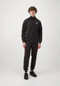 Črno športno trenirko iz mehke tkanine, sestavljeno iz jopice z zadrgo in visokim ovratnikom ter ujemajočih hlač; na prsih in nogi je logotip.