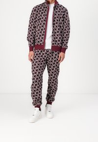 Chándal en color burdeos con un patrón geométrico, que incluye una chaqueta con cierre de cremallera y puños elásticos, combinado con joggers a juego y zapatillas blancas.