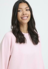 Sudadera de color rosa claro con cuello redondo, textura suave y hombros caídos. El modelo muestra un ajuste relajado y cabello oscuro ligeramente ondulado.