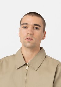 Beige, zip-up jakke med bred krage, laget av robust stoff. Gullglidelåsdetalj ved halslinjen, minimalistisk design. Jevn tekstur.