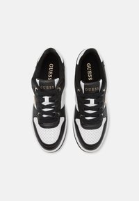 Paire de baskets Guess noires et blanches avec des bouts perforés et des lacets noirs, vues de dessus sur fond blanc.