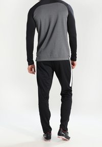 Conjunto atlético que incluye una camiseta de manga larga gris oscuro con mangas negras y pantalones negros con una franja lateral blanca, fabricado con un material flexible.